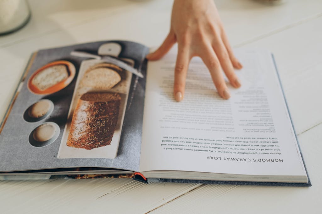 Close-up of a hand on an open clean eating cookbook with a beginner recipe page, on a white surface.